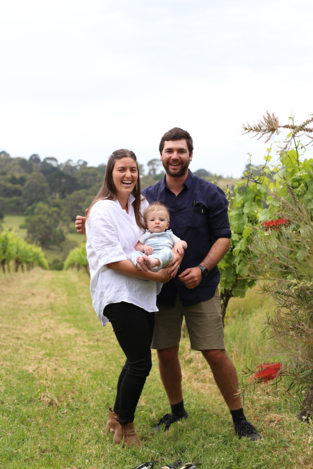 A smiling couple holding a baby, standing in a vineyard with lush greenery in the background.