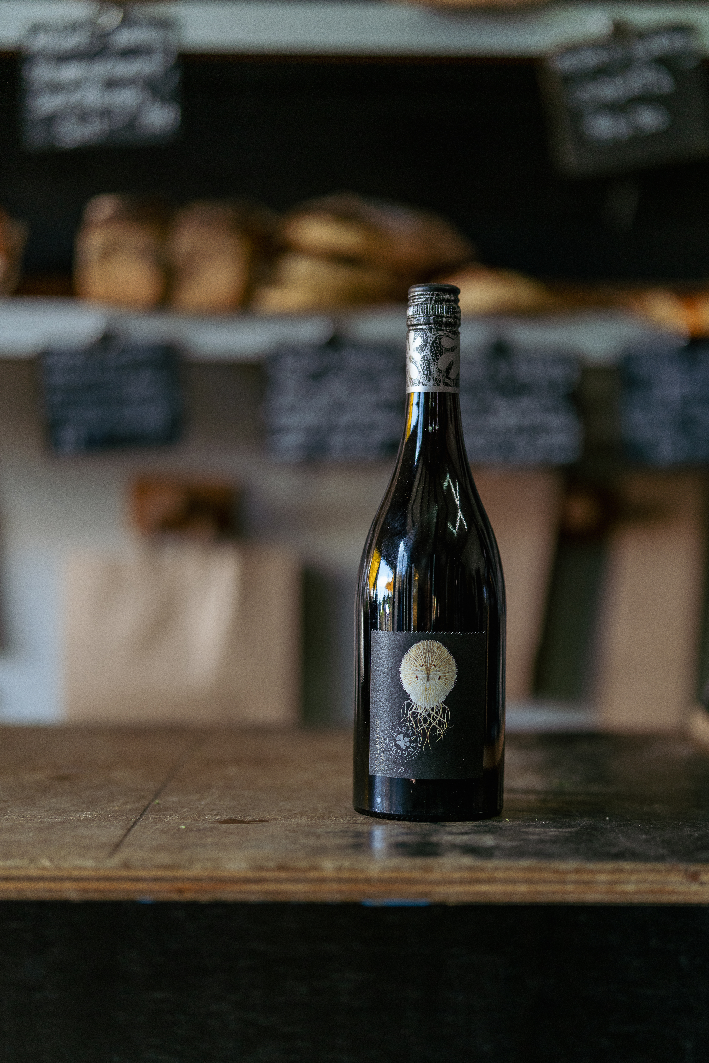 A bottle of red wine with a unique label featuring an illustration of a dandelion and roots, displayed on a wooden surface with blurred items in the background.